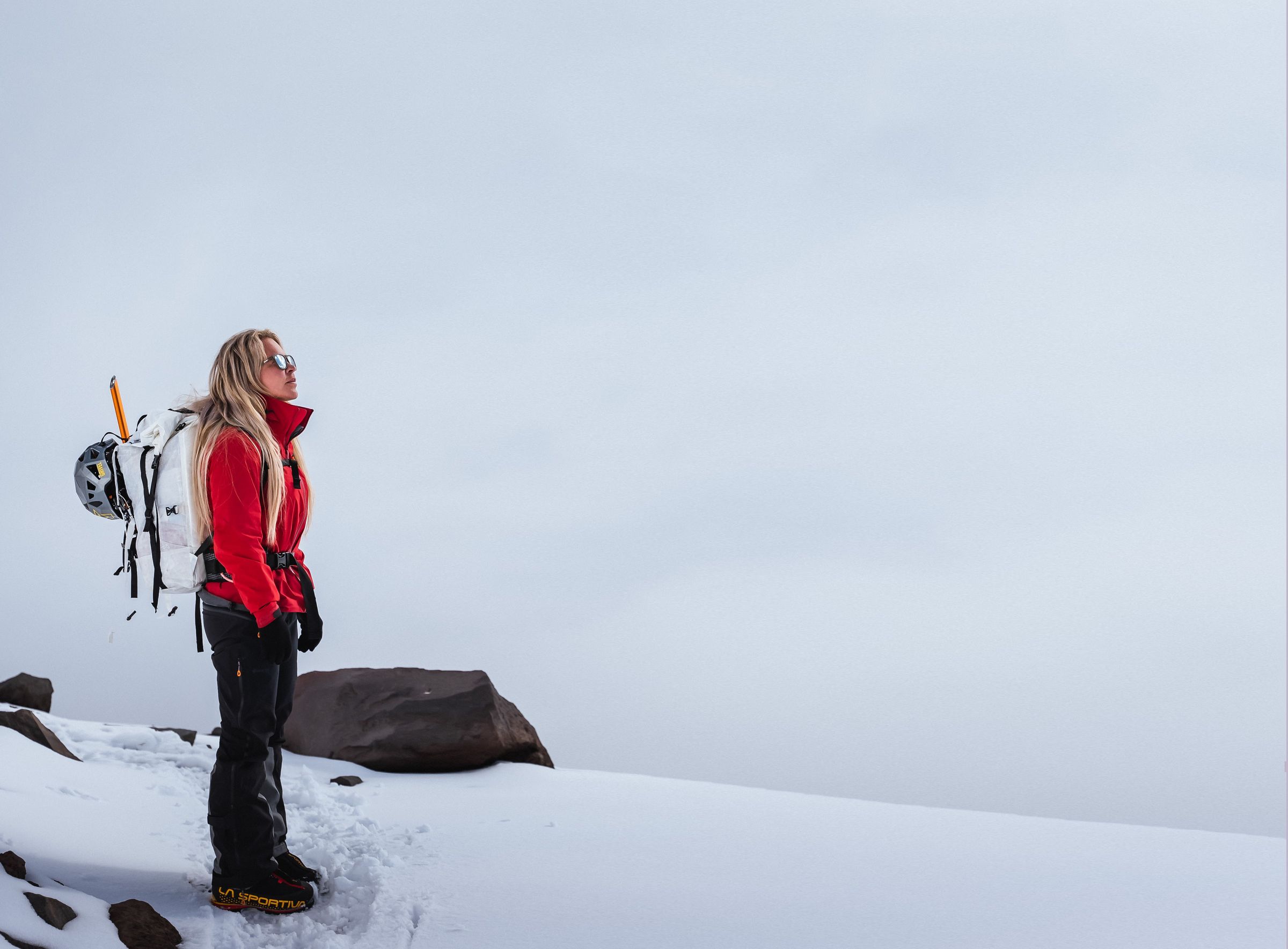 Jenn Drummond in a red jacket standing on a snowy summit ridge